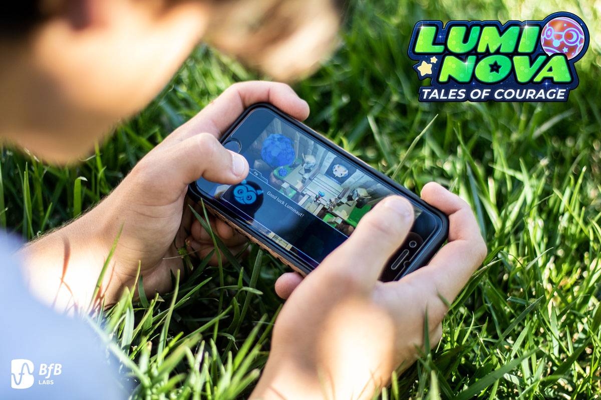 A close-up shot shows a person holding a smartphone horizontally, actively playing a game while sitting in green grass. The game screen displays colourful graphics, and the 
